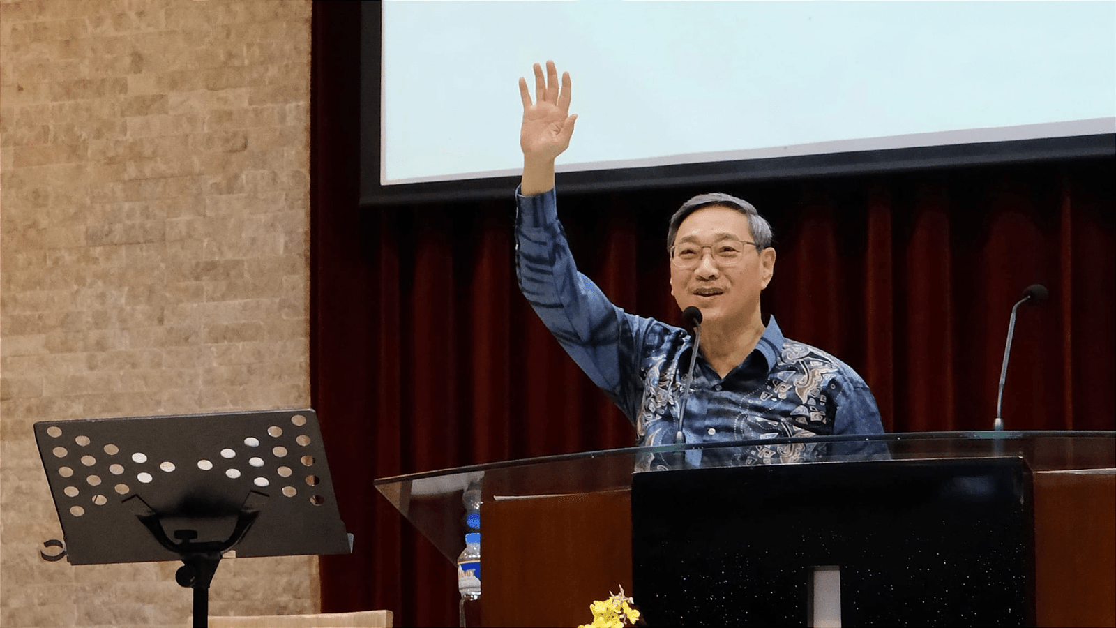 A man in a patterned shirt waving from behind a podium in a conference hall, with a microphone and a floral arrangement visible on the podium.