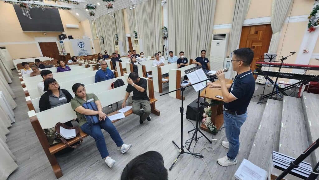 A man standing in front of a microphone in a room with people sitting on benches.