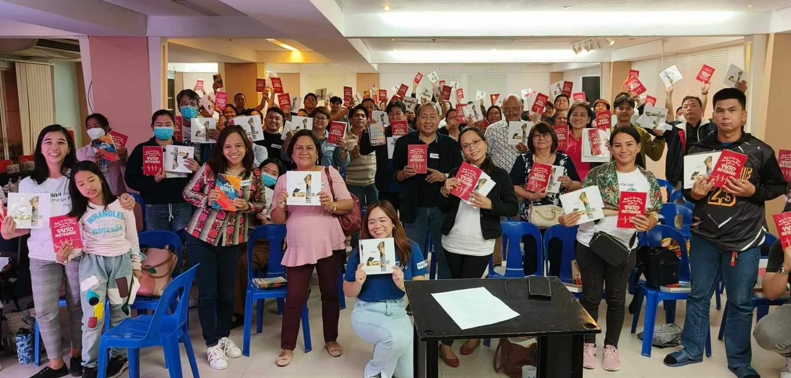 A group of people holding up books and smiling in a seminar room with chairs and a front table.