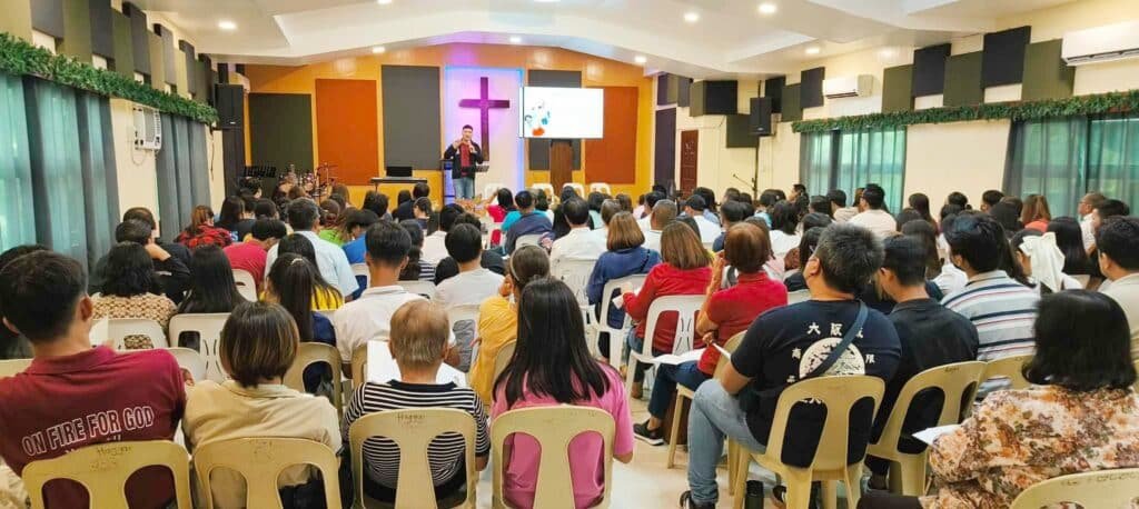 A speaker addresses a crowded room of seated attendees in a church hall, with a cross and christian imagery in the background.