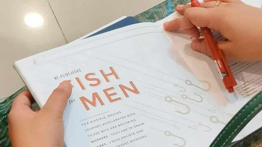 A person highlighting text in a book titled "of fish and men" with a red marker, resting on a green table.