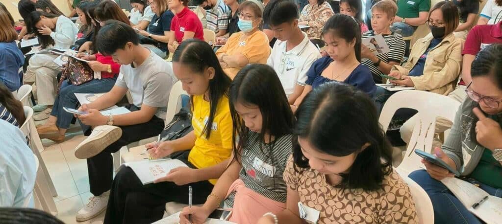 A group of people seated in a hall, concentrated on filling out forms or answering questions during an event.