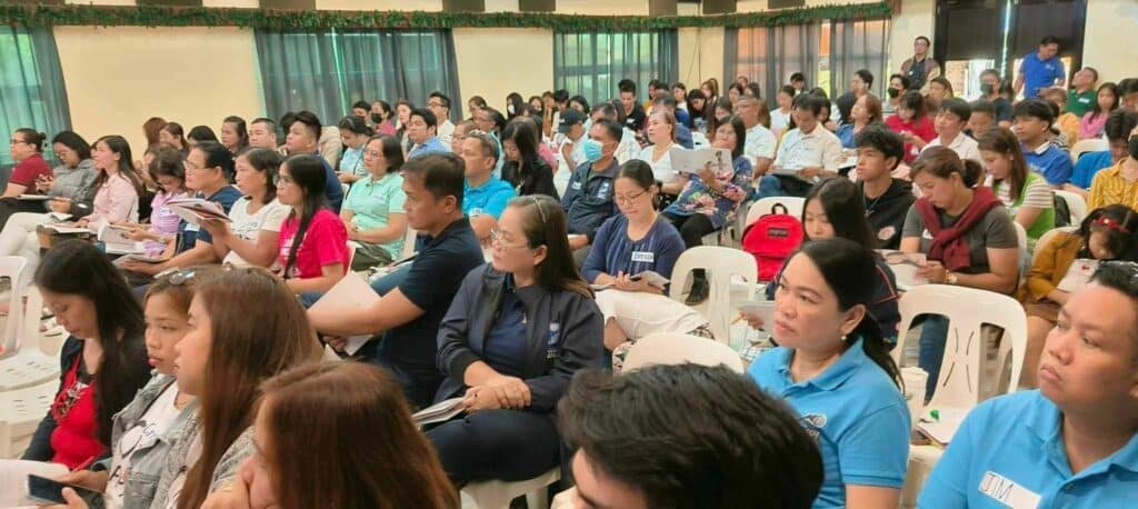 A crowded indoor meeting with people of various ages sitting on chairs, many focused on an event out of frame, some looking around.