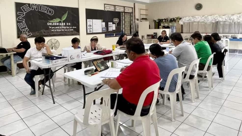 Adults sitting at tables in a classroom setting, focusing on laptops and papers, with a blackboard in the background displaying colossians 3:14.