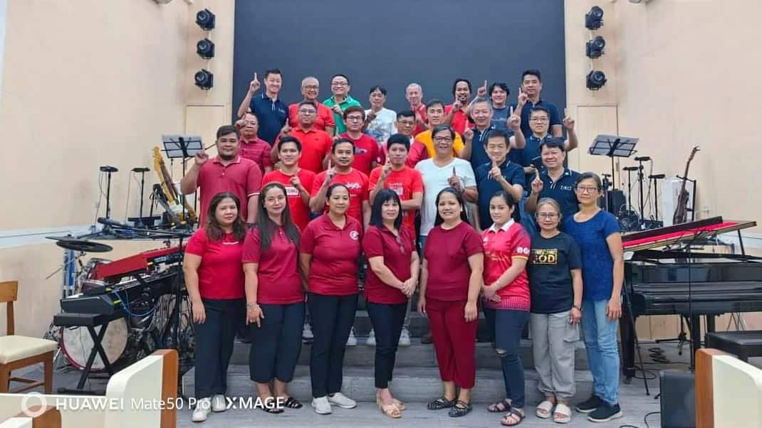 A large group of people of various ages, wearing red and blue shirts, posing for a photo in front of musical instruments on a stage.