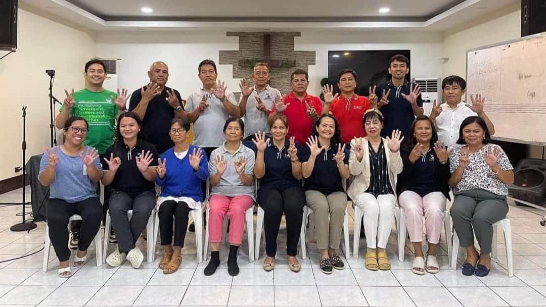 Group of fifteen adults posing in a room, showing hand signs, smiling at the camera. five are standing, ten are seated.