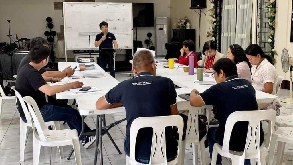 A professional workshop scene with one person lecturing and six adults sitting around tables listening and taking notes.