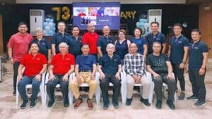 Group of fifteen people posing for a photo at an anniversary event with a banner and balloons in the background.