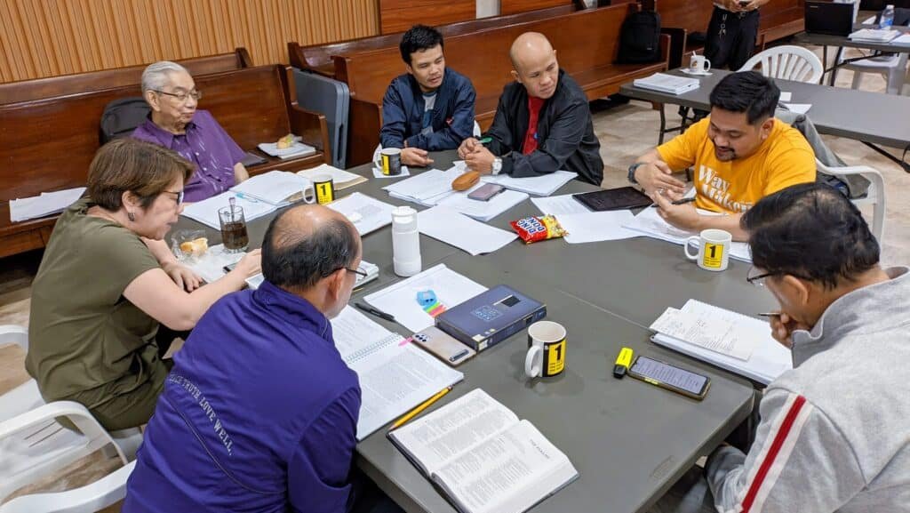 A diverse group of adults sitting around a table, engaged in discussion with books and papers in front of them in a conference room.