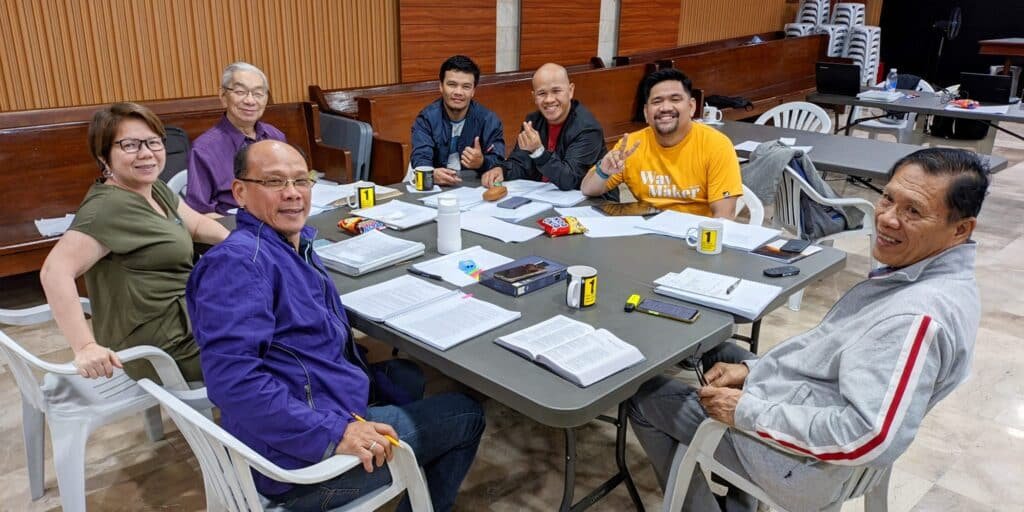 A mixed group of seven adults smiling around a table with open books and drinks, apparently in a meeting or study session.