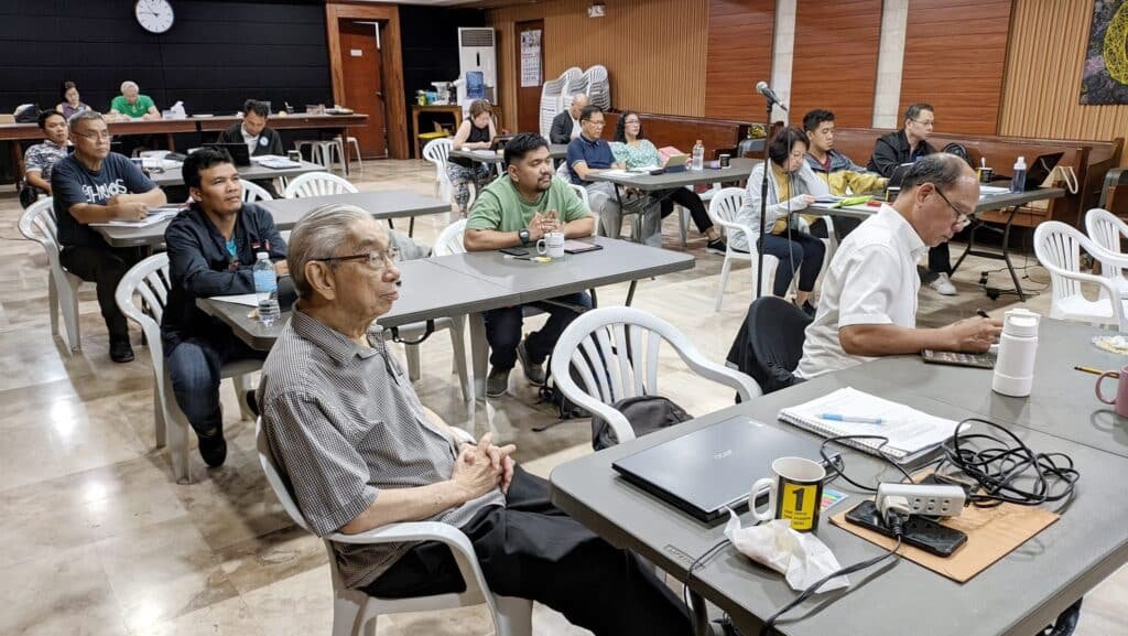 People seated at tables in a conference room, focusing on a presentation with coffee cups, papers, and electronic devices scattered around.