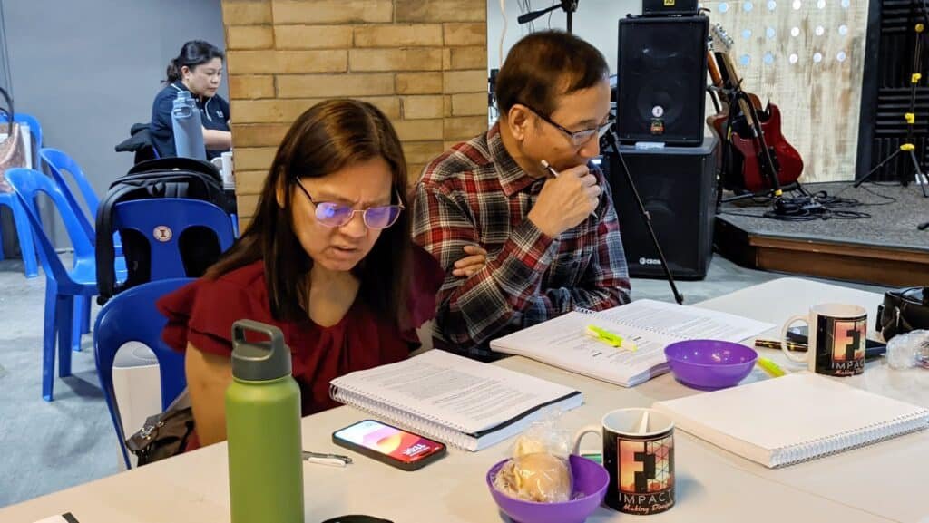 Two adults at a table with notebooks, mugs, and phones, contemplating notes, with musical equipment in the background.