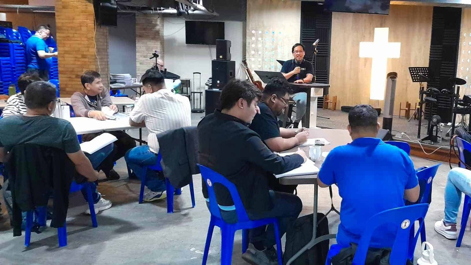 Adults seated at tables in a workshop setting inside a room with religious symbols, while a speaker addresses from the front.