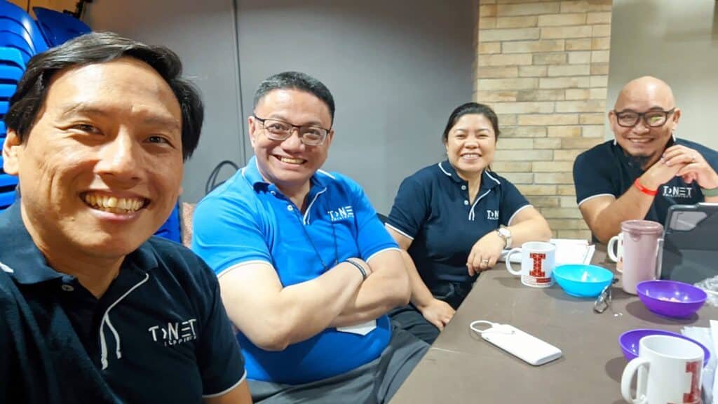 Four people, wearing blue shirts with a logo, sit at a table with mugs and a phone, smiling at the camera in a casual indoor setting.
