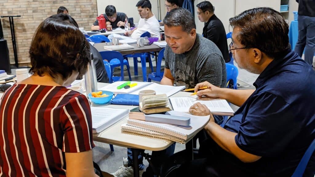 Adult students engaged in group study at a library table, discussing over notebooks and textbooks.