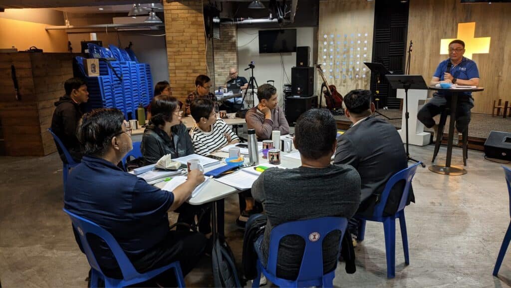 Adults seated at tables listening to a presenter in a casual workshop setting with musical instruments in the background.
