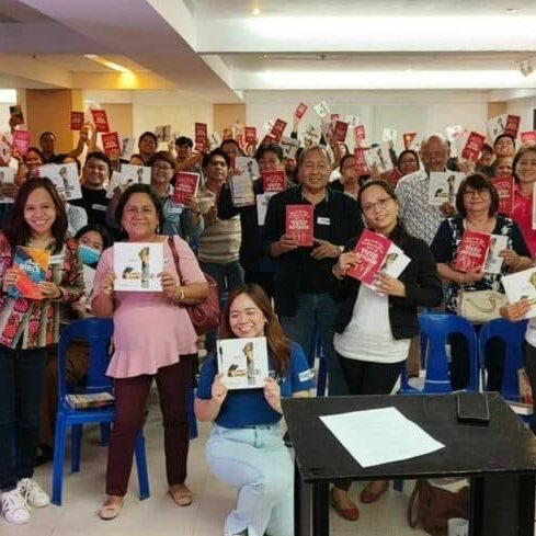 A group of people holding up books and smiling in a seminar room with chairs and a front table.