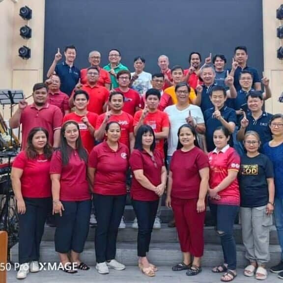 A large group of people of various ages, wearing red and blue shirts, posing for a photo in front of musical instruments on a stage.