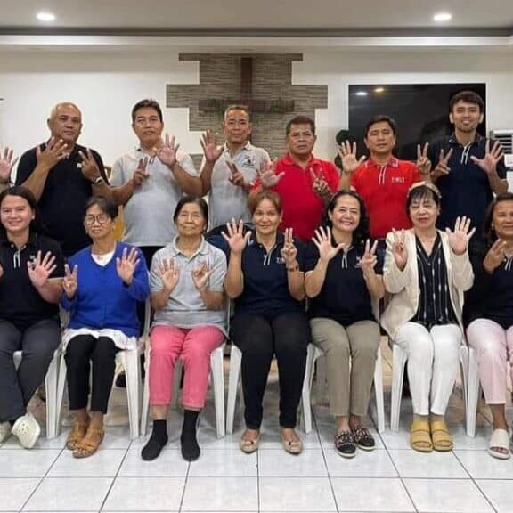 Group of fifteen adults posing in a room, showing hand signs, smiling at the camera. five are standing, ten are seated.