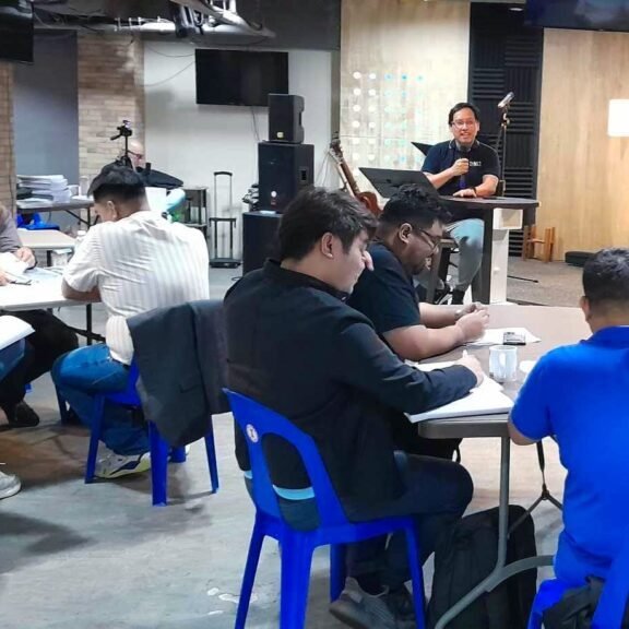 Adults seated at tables in a workshop setting inside a room with religious symbols, while a speaker addresses from the front.